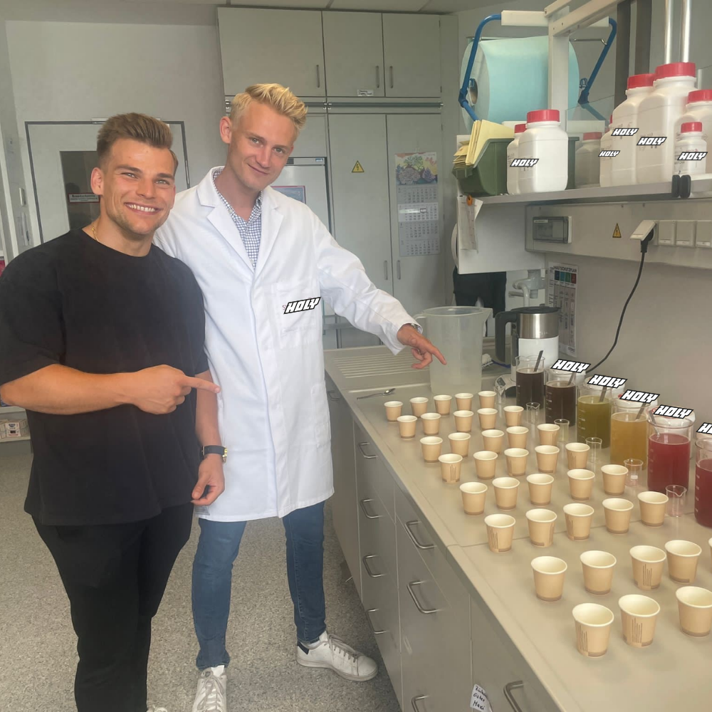 Zwei Männer stehen in einem Labor, einer trägt einen Kittel, mit vielen Pappdosen und Flüssigkeiten auf einem Tisch.