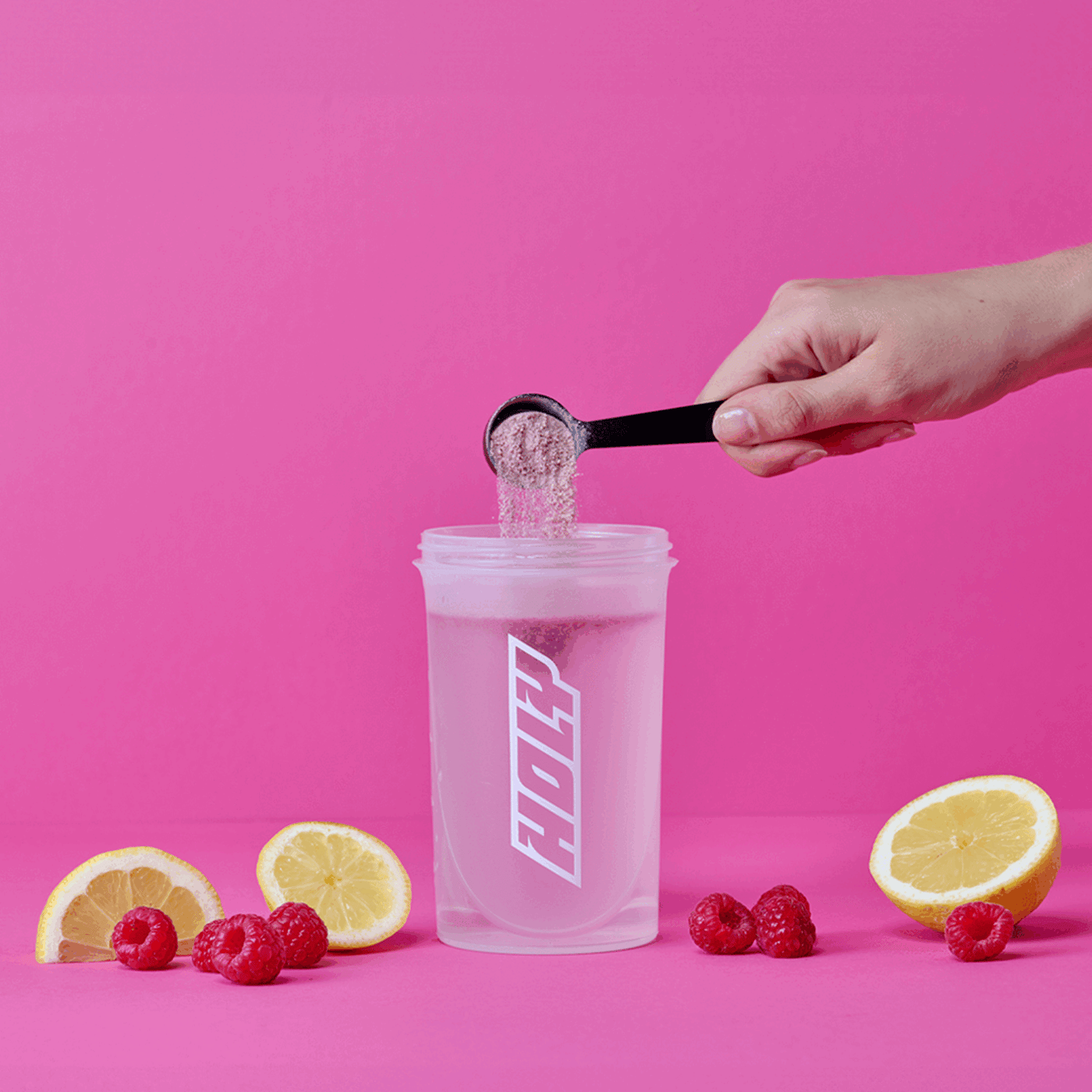 Hand fügt Pulver in einen Shaker ein, umgeben von Zitronenscheiben und Himbeeren vor einem pinken Hintergrund.