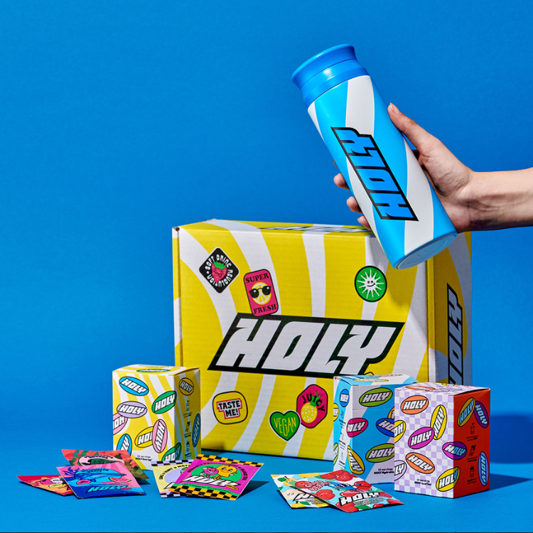 Bunte Verpackungen und Probepackungen neben einem blauen Shaker mit einer Hand vor blauem Hintergrund.