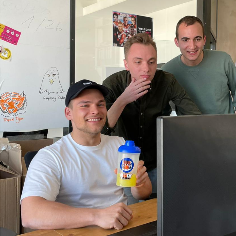 Drei Männer stehen hinter einem Schreibtisch; einer hält einen Shaker und lächelt in die Kamera.