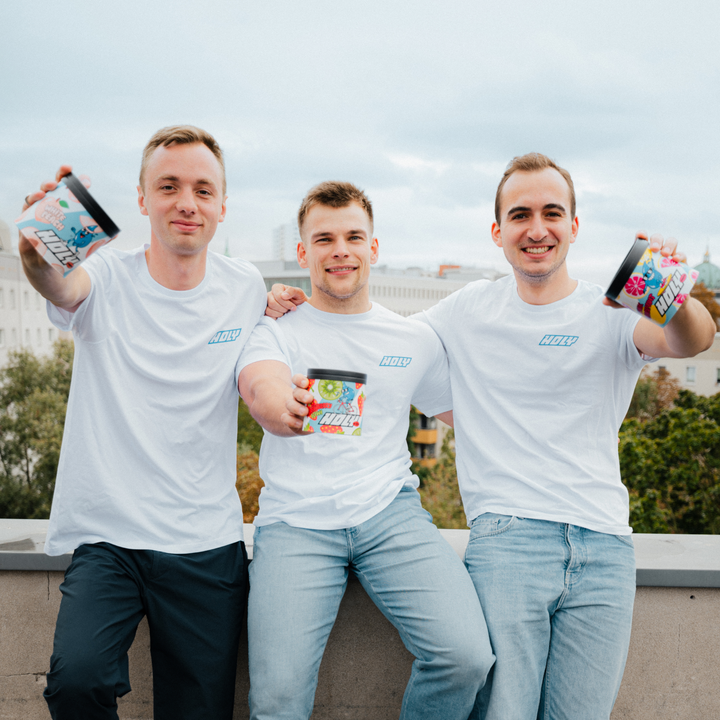 Drei Männer in weißen T-Shirts, die Pappdosen hochhalten, stehen draußen auf einem Dach vor einem bewölkten Himmel.