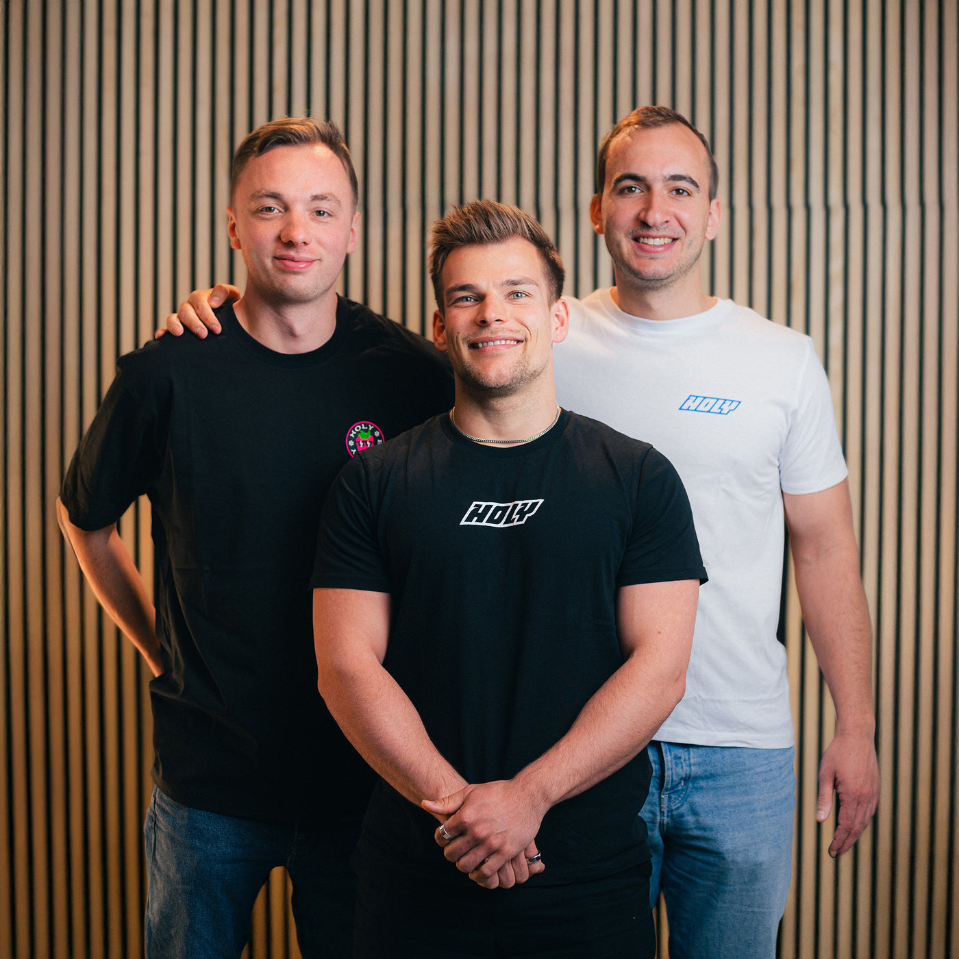 Drei Männer in schwarzen und weißen T-Shirts stehen lächelnd vor einem gestreiften Hintergrund.