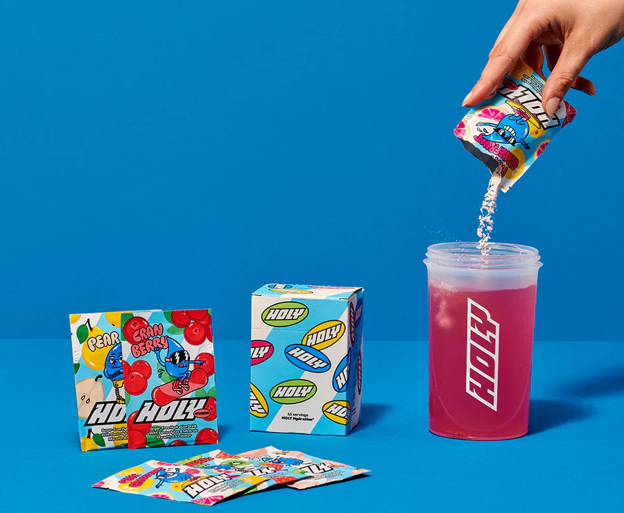 Eine Hand schüttet den Inhalt einer Probepackung in einen Shaker, daneben Pappboxen und Sachets auf blauem Hintergrund.