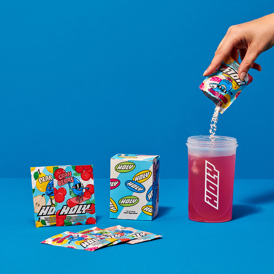 Eine Hand füllt Pulver aus einem Probepackung in einen Shaker, umgeben von weiteren Verpackungen auf blauem Hintergrund.