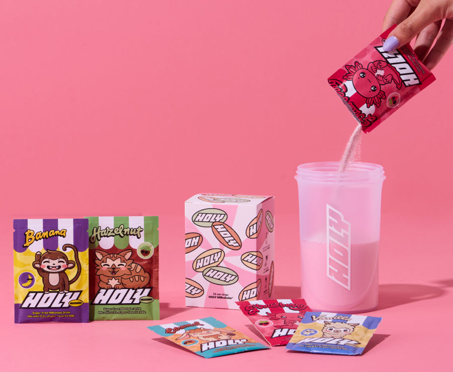 Mehrere bunte Probepackungen mit einem Karton und einem Shaker, in den eine Probepackung gegossen wird, vor rosa Hintergrund.