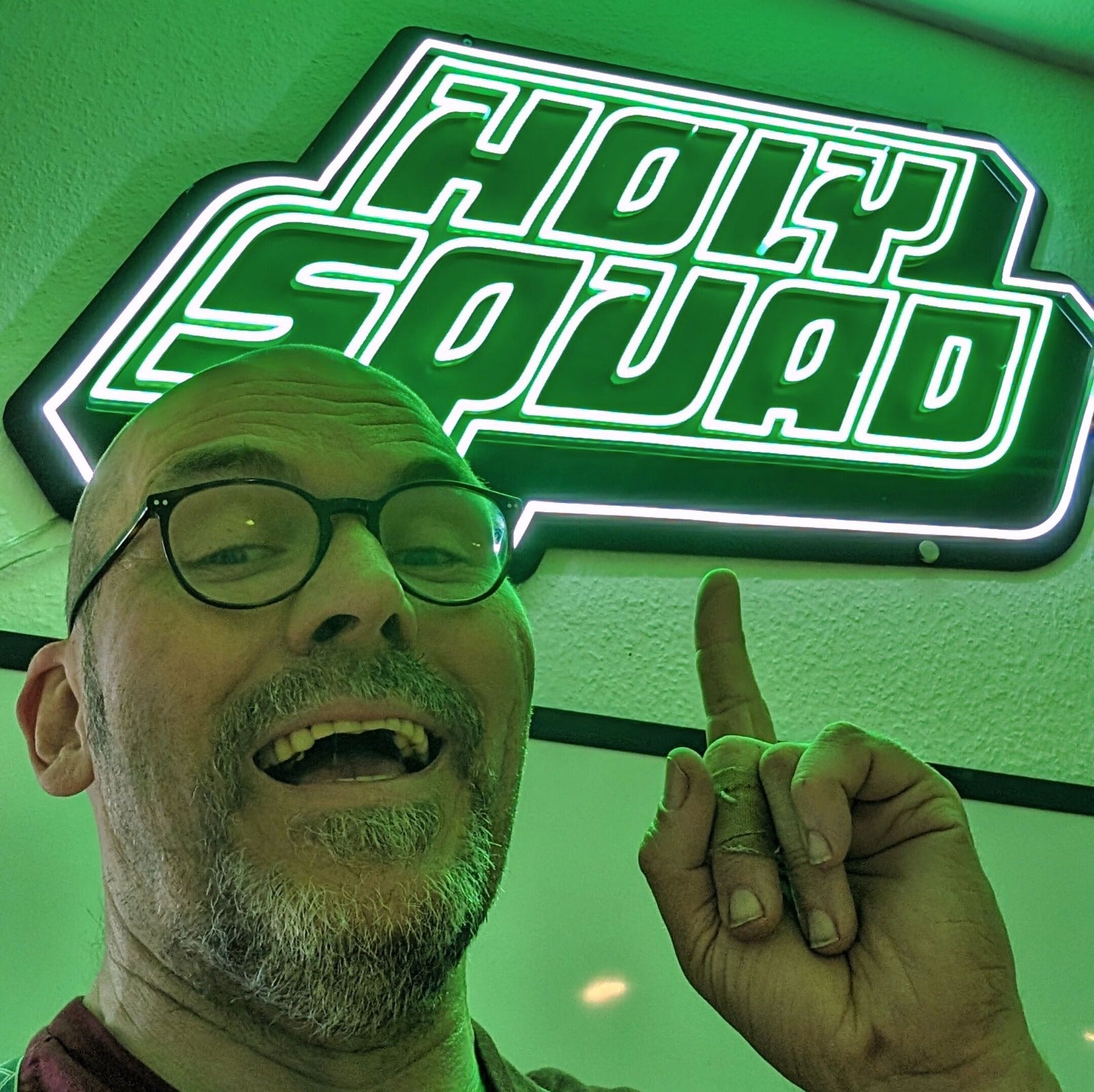 Person mit Brille zeigt lächelnd auf ein grünes Neonschild mit der Aufschrift "HOLY SQUAD".
