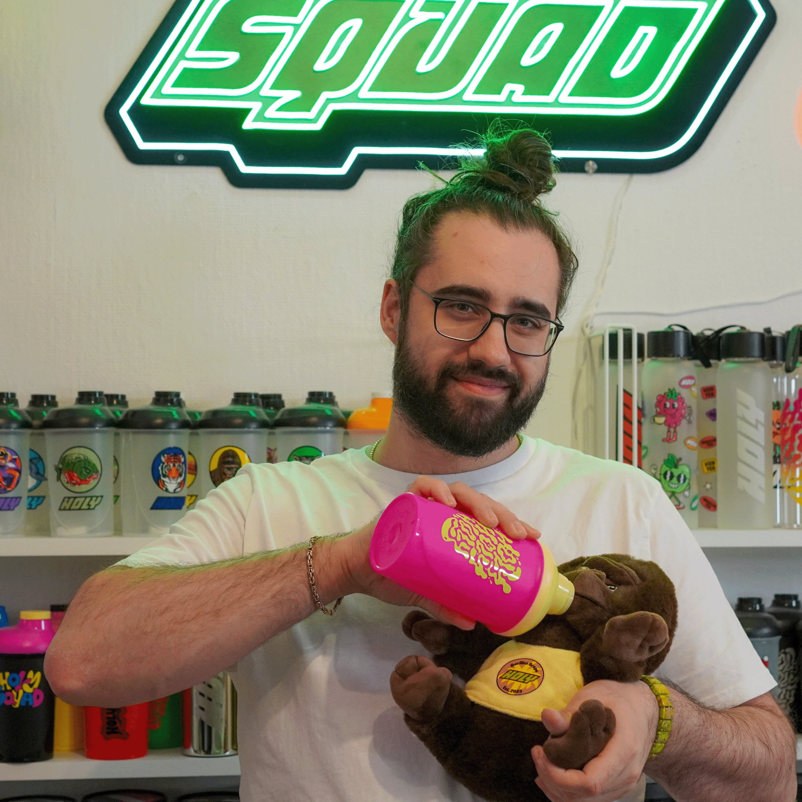 Ein Mann mit Brille füttert ein Plüschtier aus einem pinken Shaker. Im Hintergrund sind weitere Shaker auf einem Regal.