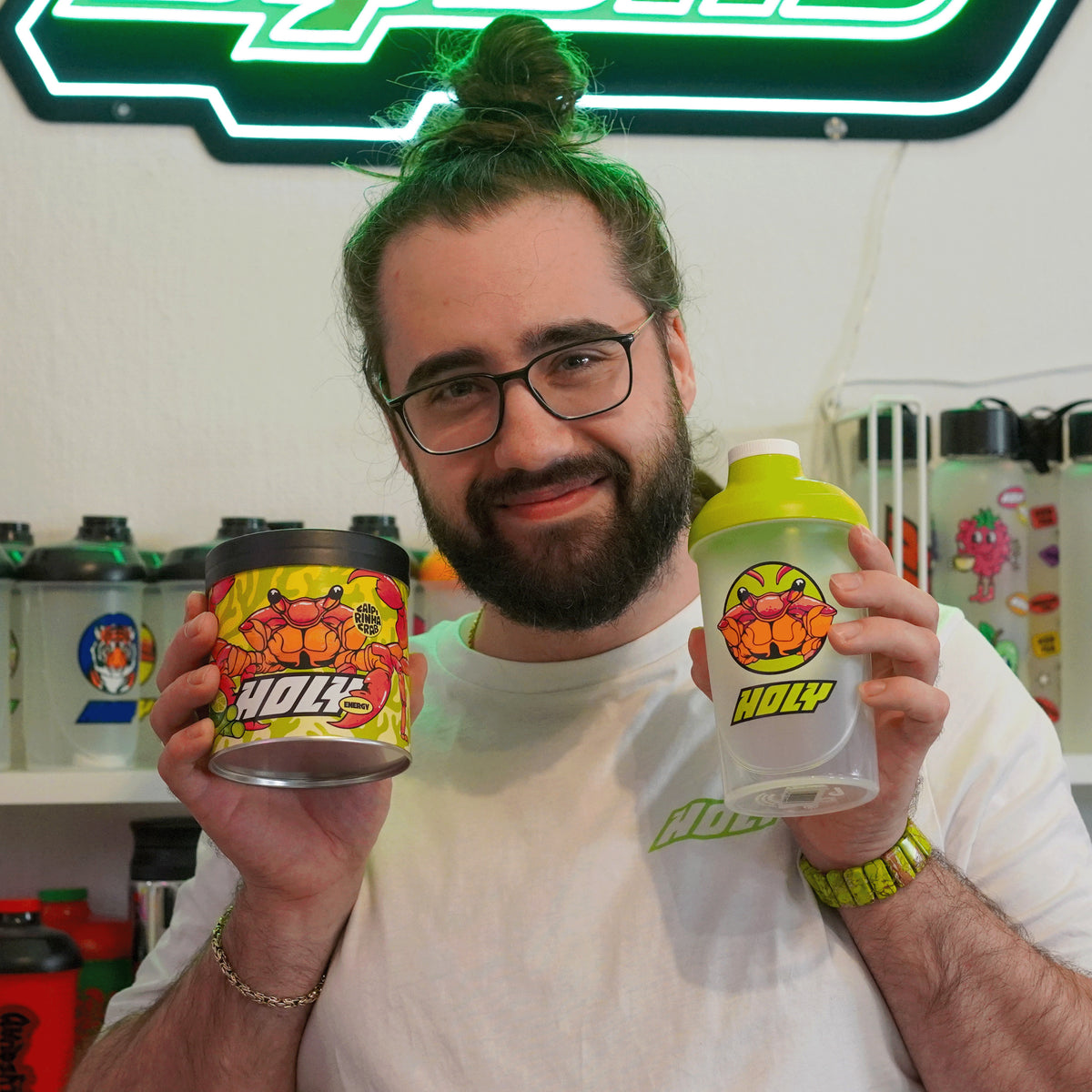 Mann hält eine bunte Pappdose und einen Shaker mit ähnlichem Design, umgeben von weiteren Flaschen im Regal.