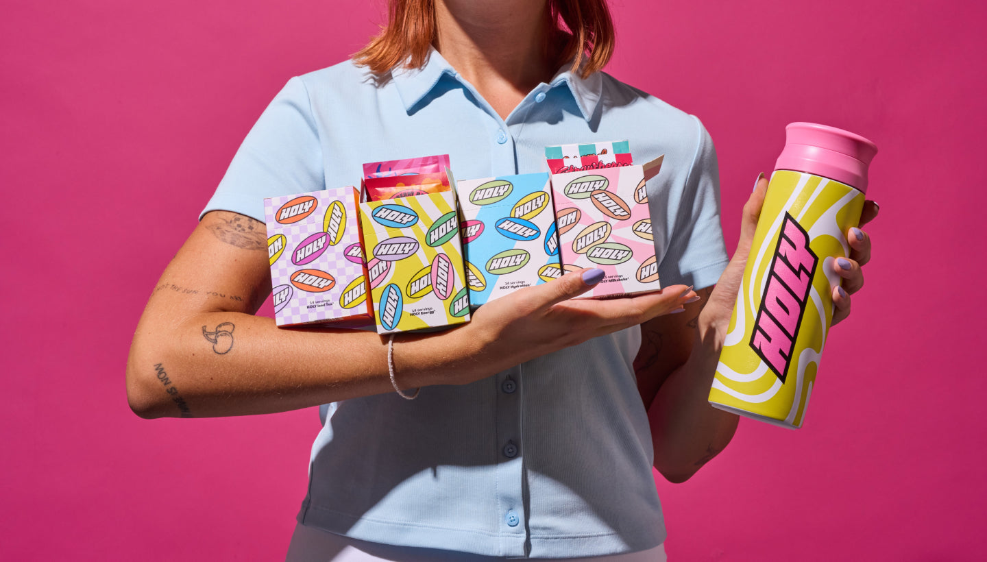 Frau hält vier bunte Pappboxen mit Probepackungen und einem gelb-weißen Shaker vor einem pinken Hintergrund.