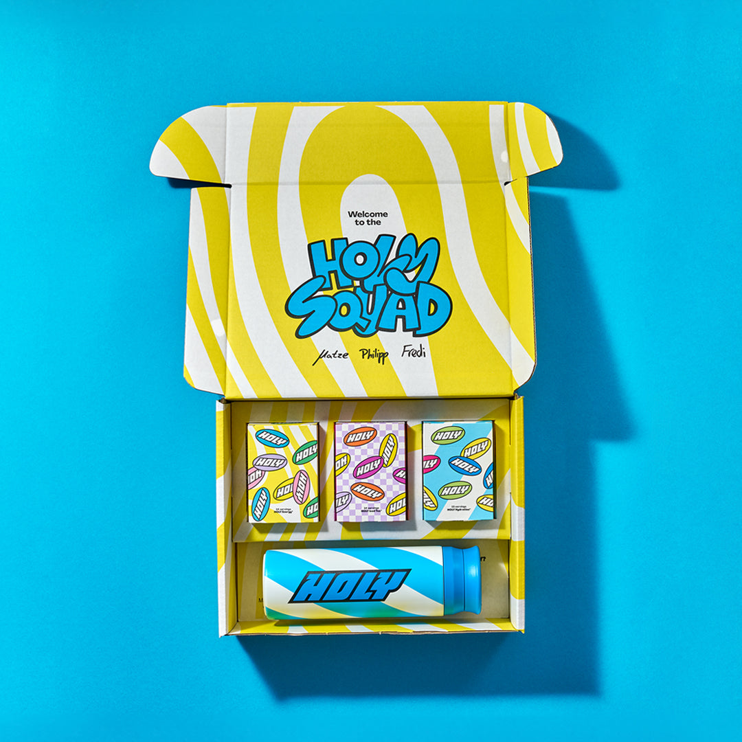 Offene Pappbox mit drei Pappdosen, einigen Probepackungen und einem Shaker, alles auf blauem Hintergrund.