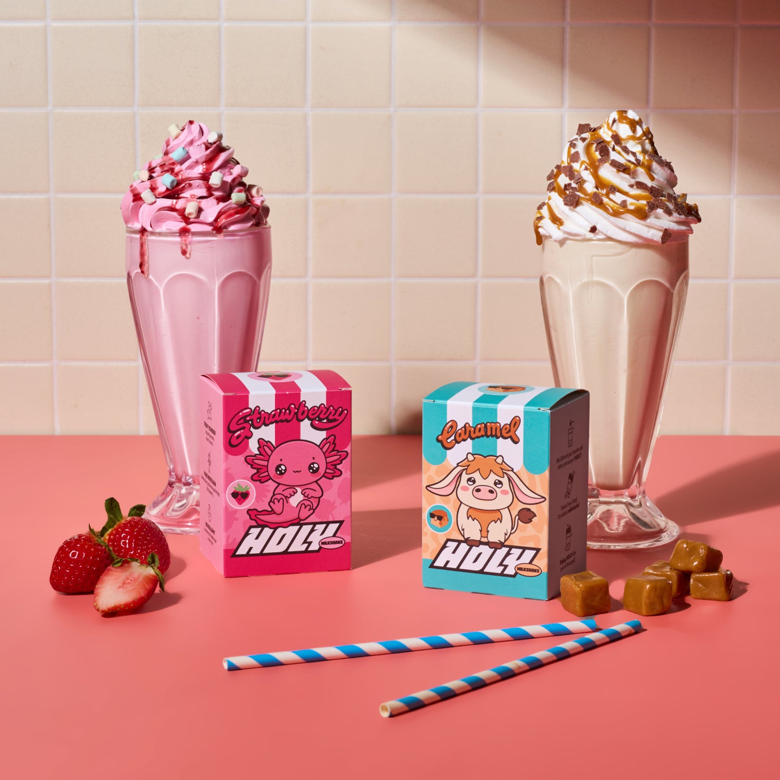 Zwei Milchshakes mit Sahne, Erdbeeren, Karamellen und zwei bunte Pappboxen auf einem rosa Tisch vor Fliesenhintergrund.