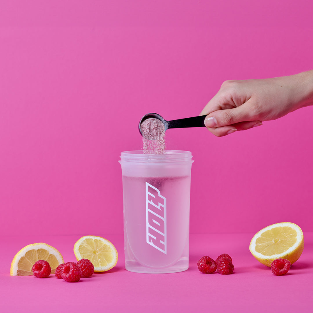 Eine Hand fügt Pulver mit einem Messlöffel in einen Shaker, umgeben von Zitronenhälften und Himbeeren, auf rosa Hintergrund.