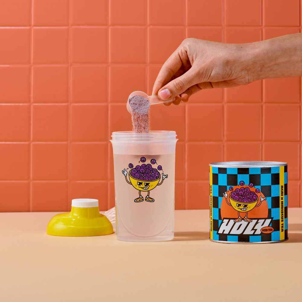 Eine Hand füllt Pulver in einen durchsichtigen Shaker mit buntem Cartoon-Aufdruck, neben einer gemusterten Pappdose.