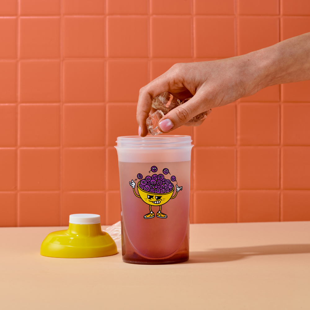 Eine Hand gibt Eiswürfel in einen halb vollen Shaker mit gelbem Deckel und fröhlicher Cartoon-Grafik.