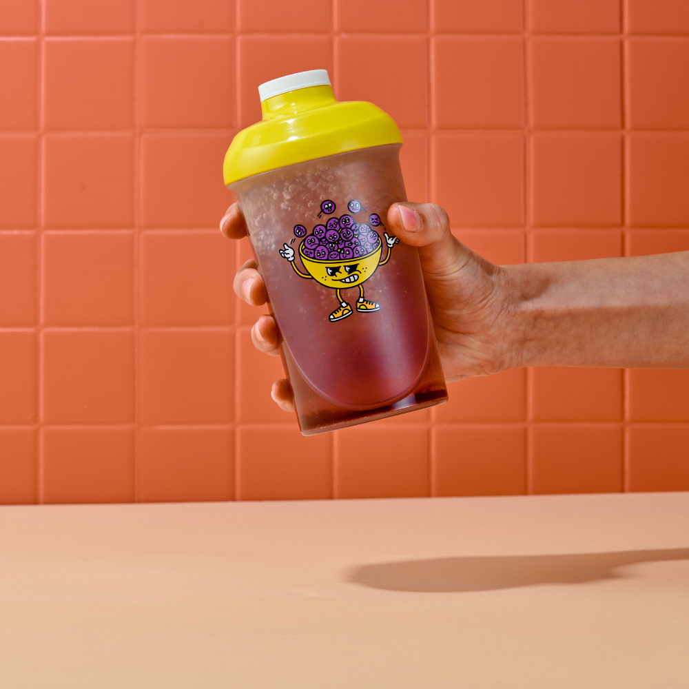 Hand hält einen Shaker mit gelbem Deckel und lustigem Cartoon-Motiv vor einer orangefarbenen Kachelwand.